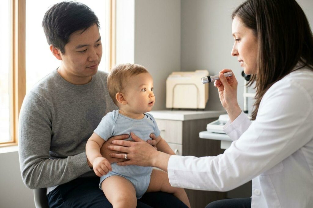 Doctor using a small light to examine a baby's eye in a clinic, while an adult holds the infant on their lap.